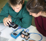 The picture shows two students experimenting at a table and using a smartphone to observe an experiment. Next to them are measuring instruments and a construction with coils used for a technical or physical investigation.