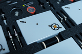 The picture shows a tool and experiment box containing various equipment and materials for school experiments. These include gray plates, clips, a compass and other utensils organized in a foam insert.