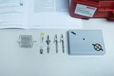 The picture shows an arrangement of experimental materials that can be used for a physics lesson. Among other things, you can see a spiral, various connecting elements and a grey case, together with a teaching manual.