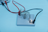 The picture shows an experimental device with a spiral spring and an LED on a gray base connected by colored wires. This arrangement is intended for illustrating electrical circuits and experiments in educational settings.