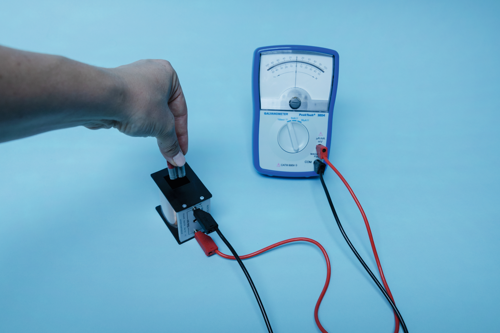 The picture shows a hand holding two magnets in a device while a galvanometer is ready to measure electrical parameters. The components are designed for physics experiments in the educational field.