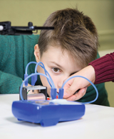 The picture shows a boy working concentratedly on an experimental device while being assisted by another person. The device is connected to colorful cables used for scientific experiments.