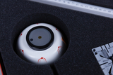 The image shows a circular object lying in a padded holder, surrounded by black foam. Red patterns can be seen on the surface of the object, while gray components for further experiments lie next to it.