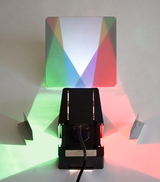 The picture shows an experimental device consisting of a light source and a prismatic surface. It produces colored light rays in red, green and blue that are projected onto the wall to demonstrate light refraction and mixing work.