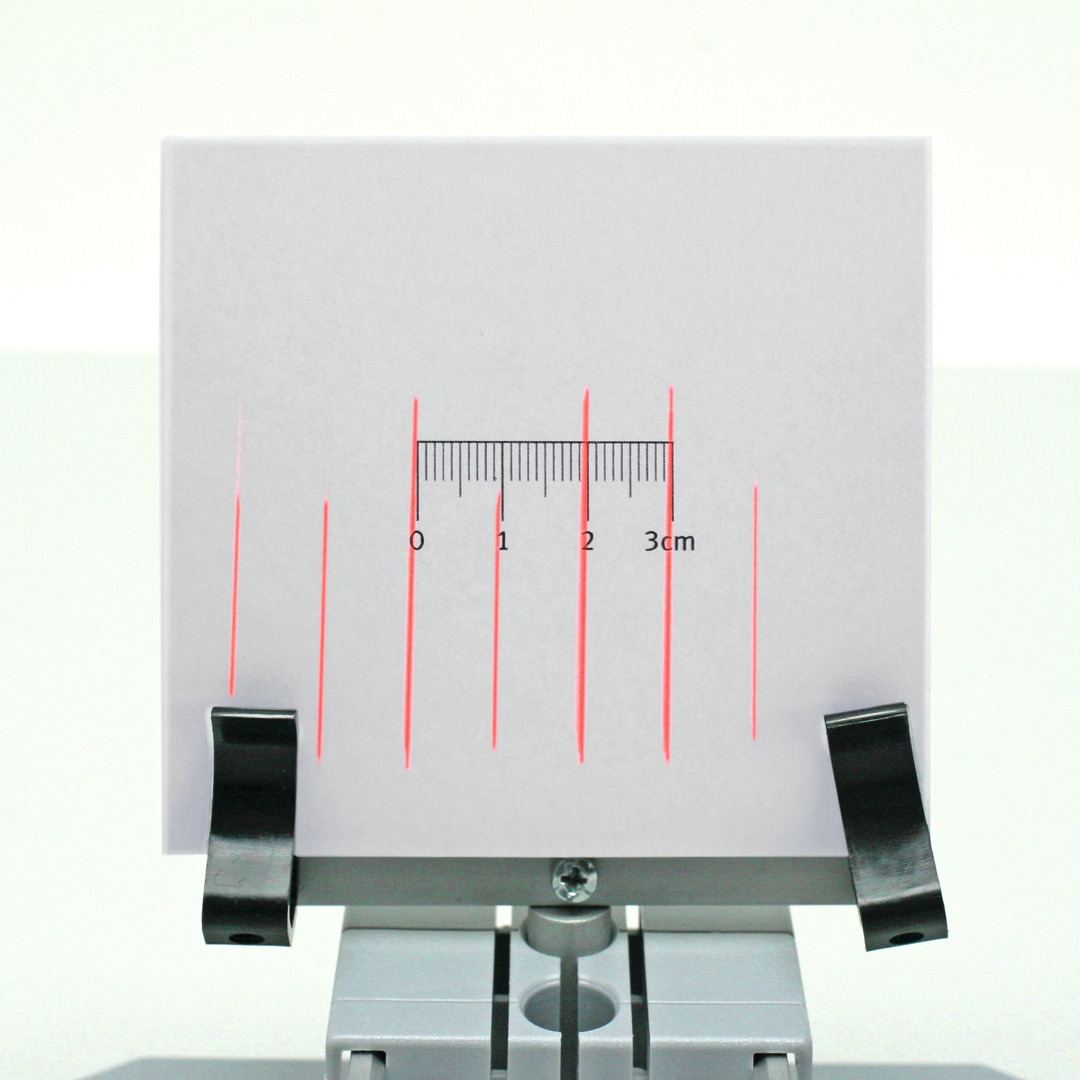 The image shows a white sheet with red parallel lines and a scale marking from 0 to 3 cm. It is attached to a holder used for educational experiments.