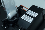 The image shows a technical device with a transparent chamber and an electronic control panel containing safety instructions in English and German. It appears to be designed for experimental purposes in education and has electrical connections.