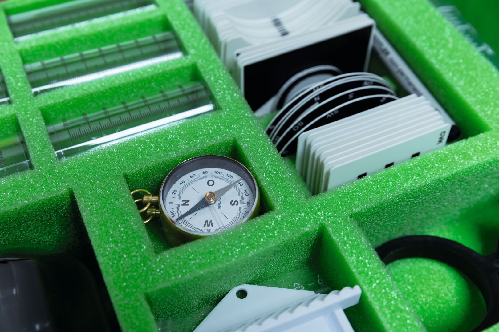 The picture shows a set of experimental materials arranged in a green foam insert. Among other things, there is a compass and various measuring devices that can be used for educational projects.