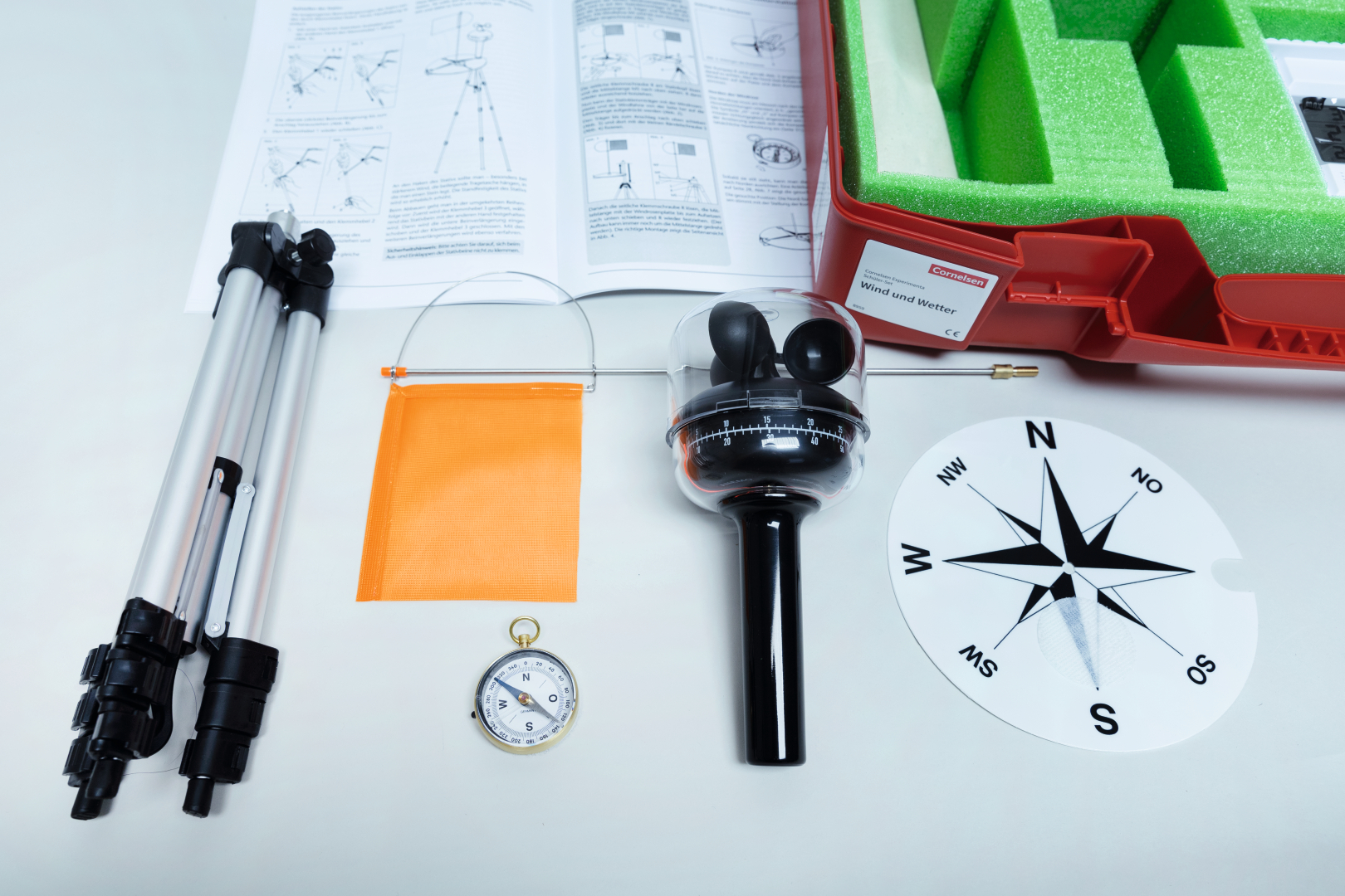 The picture shows a collection of experiment materials for teaching, including a compass, a wind vane, a tripod and instructions. These tools are packed in a red case and are used to study wind and weather.