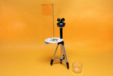 The picture shows an experimental device that stands on a tripod and contains a compass disc and a small flag. Next to the device is a transparent container, presumably for conducting experiments in the educational field.
