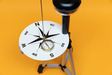 The image shows an experimental device with a large compass rose and a pendulum mounted on a sturdy tripod. The background is bright orange, which draws attention to the device and makes it ideal for teaching and learning purposes in education.