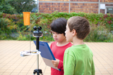 The picture shows two children working at an experiment stand with a weather instrument. One of the children is holding a clipboard and looking at the instruments while the other is watching attentively.