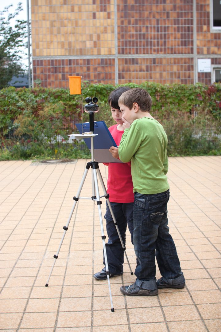 The picture shows two children working together on an experimental device mounted on a tripod. One child is holding a block in his hand while the other is carefully watching what they are doing with the device.