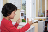 The image shows a boy curiously looking at a thermometer on a window. In the background are typical school supplies and a wall with a calendar, indicating an educational environment.