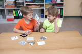 In the picture, two children are sitting at a table, looking at cards with different patterns and shapes. They seem to be actively discussing the cards and enjoying their experimental game.