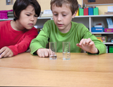 The picture shows two children concentrating on two measuring cups of water in front of them. One of the children is wearing a red shirt, the other a green one, while they discuss the fill levels of the cups.