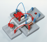 The picture shows an experimental device consisting of several gray base elements connected to each other. On the plates there is a battery unit, a switch and a copper coil used for electrical experiments in education.