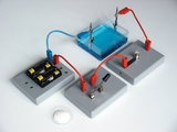 The picture shows an experimental setup with an electrochemical cell, consisting of a water layer with blue liquid and various connections. Batteries, connecting wires and measuring instruments for conducting experiments are visible.