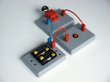 The picture shows an experiment set consisting of several gray plates, cables, a metallic ball and a button. It is used to illustrate electrical circuits and can be used in education for practical exercises.