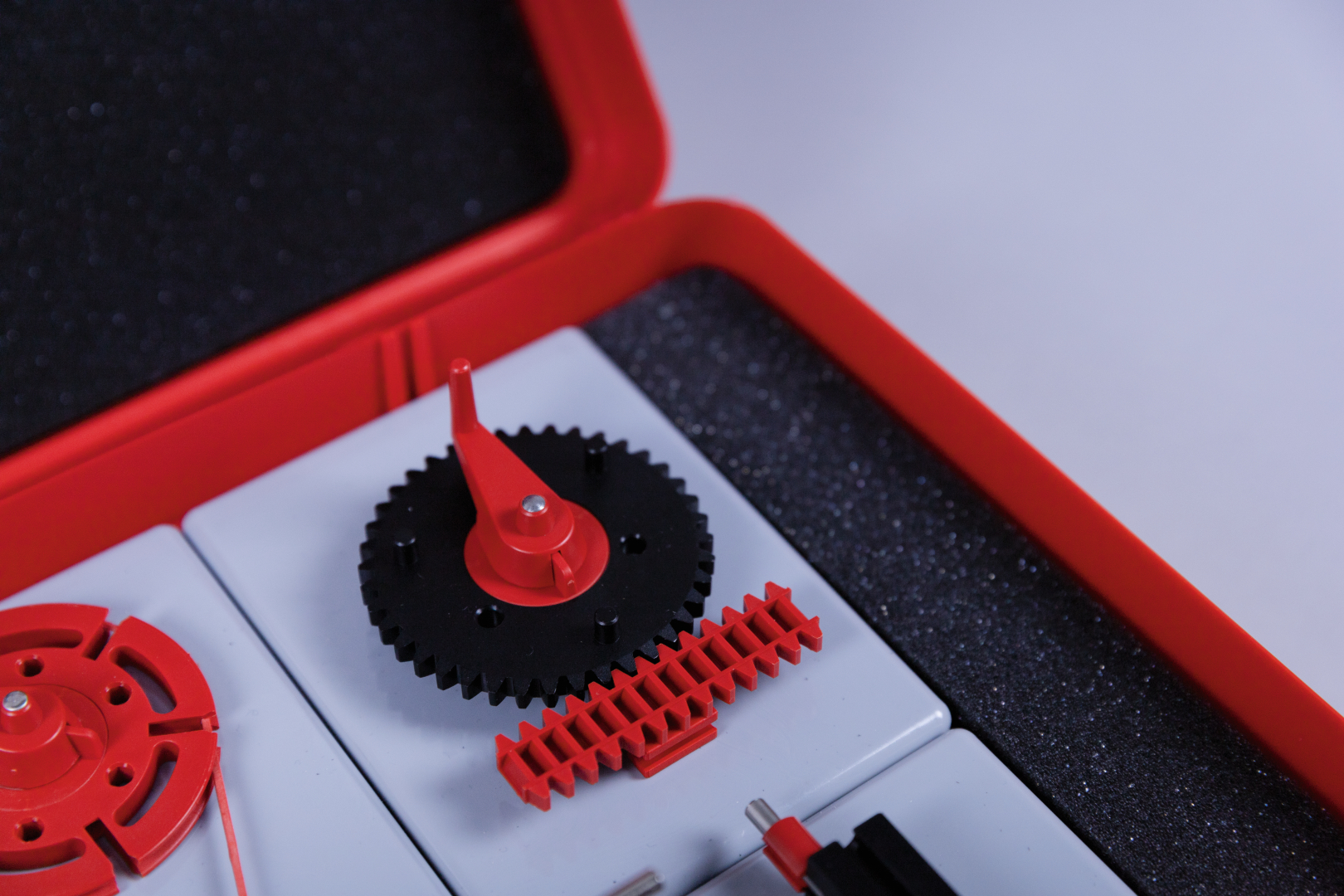 The picture shows an experiment kit with various mechanical components, including gears and a red control component. The parts are neatly arranged in a red case that is lined with foam to protect them.