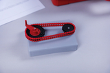 The image shows a mechanical model with a red chain drive and two black gears, placed on a gray platform. It is probably intended to illustrate principles of mechanics and technology in educational settings.