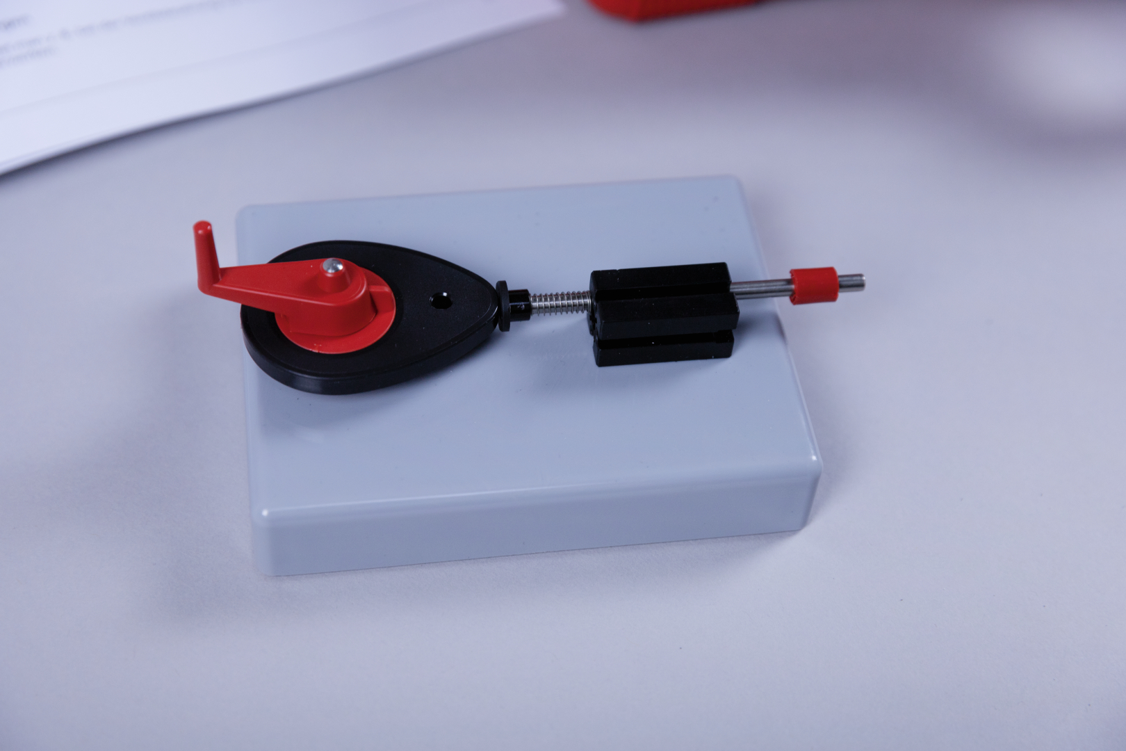 The picture shows an experimental device with a red lever and a black housing mounted on a grey base. It seems to be designed for various scientific experiments in education.