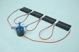 The picture shows an experimental set with several solar panels connected to a small electric motor by red cables. The motor has a blue propeller that is in motion, demonstrating the conversion of solar energy into mechanical energy.