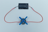 The picture shows a simple experimental setup consisting of a small motor with blue propellers connected to a solar cell module. This arrangement demonstrates how sunlight can be converted into mechanical energy.