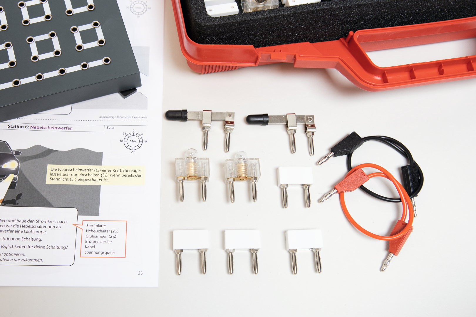 The picture shows various electronic components, including switches, light bulbs and connecting cables, used for educational experiments. Alongside the materials is an instruction manual with information on how to conduct an experiment to control fog lights.