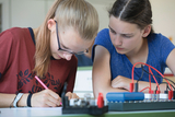 The picture shows two schoolgirls working concentratedly on an experimental project. One of the girls is drawing something on paper while the other is carefully looking at the materials on the table.