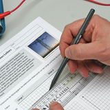 The image shows a hand writing with a pencil on a worksheet with a diagram and graphic representation of high voltage power lines. The worksheet appears to contain information about the electrical resistance of conductors.