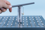 The image shows a hand holding a metal rod above a silver cylinder that is resting on a plate with electrical contacts. A small body is hanging from the cylinder, indicating an experiment to demonstrate physical principles.