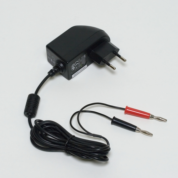 The picture shows a black power supply with a Euro plug and two cables with crocodile clips in red and black. It is designed for use in educational experiments and provides a connection for electrical applications.