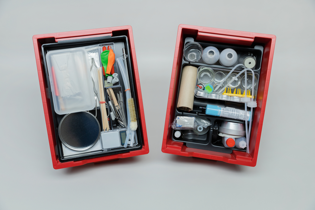The picture shows two red containers filled with various experimental materials. The boxes contain utensils such as measuring instruments, glasses, tubes and craft materials that are suitable for scientific experiments in educational settings.