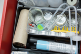 The image shows a collection of laboratory equipment and materials arranged in a red container. These include glass containers, pipettes, a cardboard cylinder and a sprayer used for educational experiments.