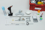 The image shows a collection of experimental supplies, including a burner, test tubes, a precision balance, and various chemicals in vials. The items are neatly arranged on a white background and are used for science education.