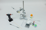 The image shows laboratory equipment used for chemical experiments, including a heater, a beaker of liquid, a digital scale, and various chemicals in vials. The arrangement of the equipment and materials suggests an experimental setting.