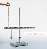 The image shows a massive lever with a beam balance and weights for conducting experiments. A hand holds a measuring device while the system is used to illustrate physical principles such as balance.