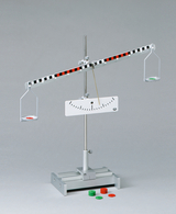 The picture shows a seesaw with two bowls attached to a horizontal beam that is supported on a central pivot point. In addition, there are colored discs in red and green on the table, which are probably used for determining weight or for experimentation.