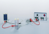 The picture shows an electrolysis experiment set with a solar module, two measuring cylinders for hydrogen and oxygen, and a control unit for regulating current and voltage. It is used to demonstrate hydrogen production through electrolytic processes.