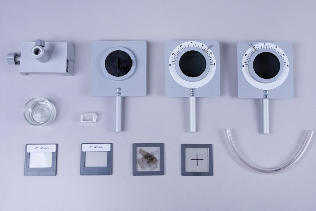 The picture shows a selection of experimental equipment, including measuring instruments such as a deflection tube and various microscope slides. In addition, a glass container and a transparent tube can be seen, which can be used for physics experiments.