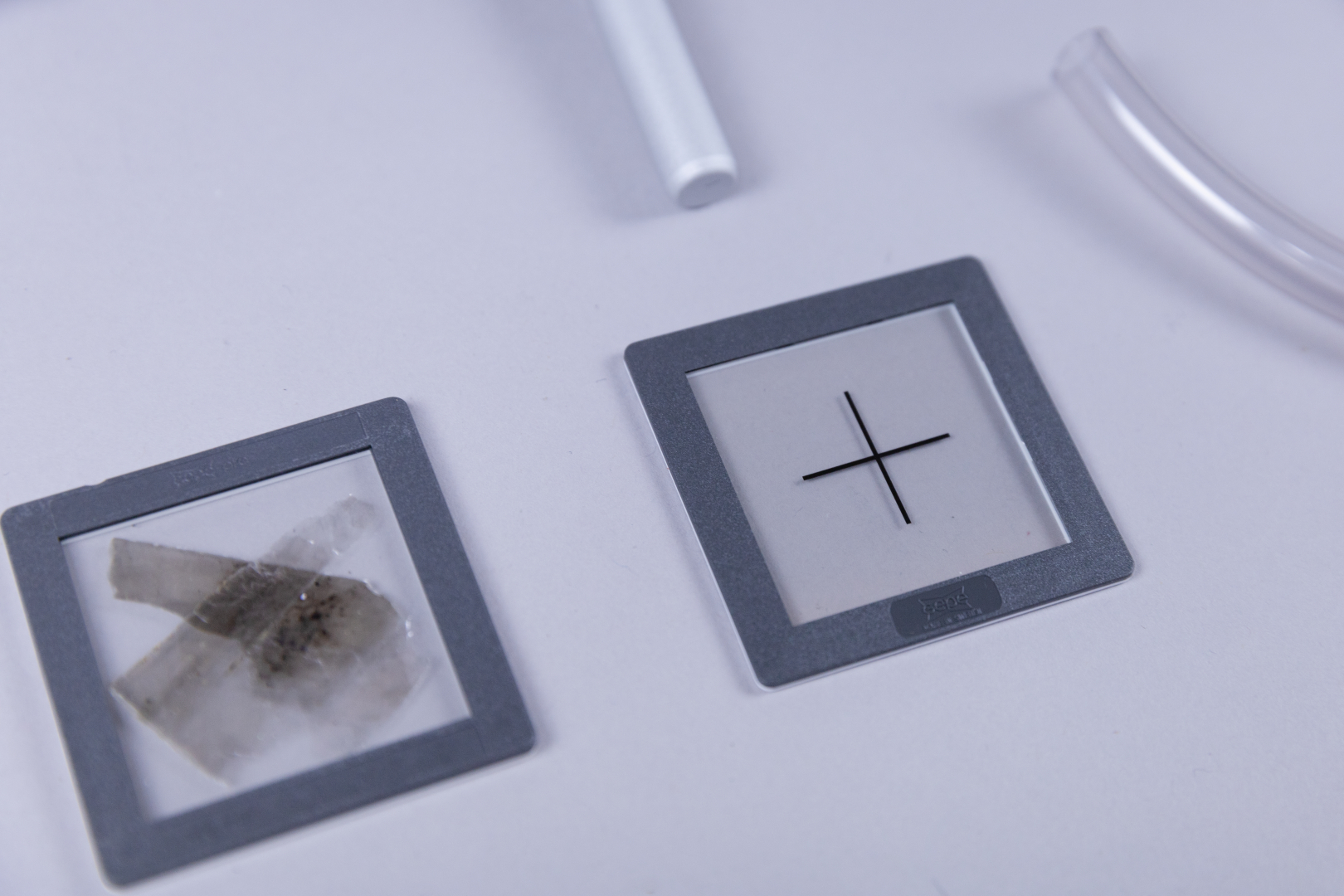 The picture shows two slides, one with various mineral samples and the other with a black cross pattern. Next to it is a transparent tube, possibly used for experimental purposes.