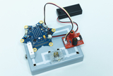 The picture shows an electronic experiment board with a central board in the shape of a star that contains various electronic components. The board is connected to a battery compartment and control elements that can be used to conduct experiments in educational settings.