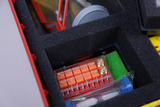 The picture shows a toolbox from an experiment supplies set that is filled with various materials. Inside you can see an orange container module, glass tubes and a green sponge, which can be used for experiments.