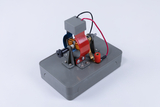 The picture shows a gray experimental device consisting of various metallic components, including a coil and electrical connections. It is probably intended to illustrate basic physical principles, such as electromagnetism or engine construction.