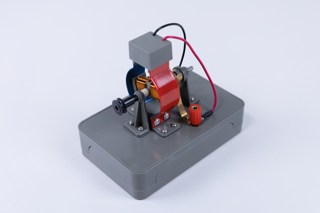 The picture shows a gray experimental device consisting of various metallic components, including a coil and electrical connections. It is probably intended to illustrate basic physical principles, such as electromagnetism or engine construction.