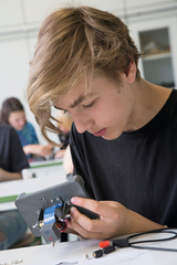 A student is working concentratedly on a technical experimental device, into which he is inserting various components. In the background, other students can be seen who are also working with experimental materials.