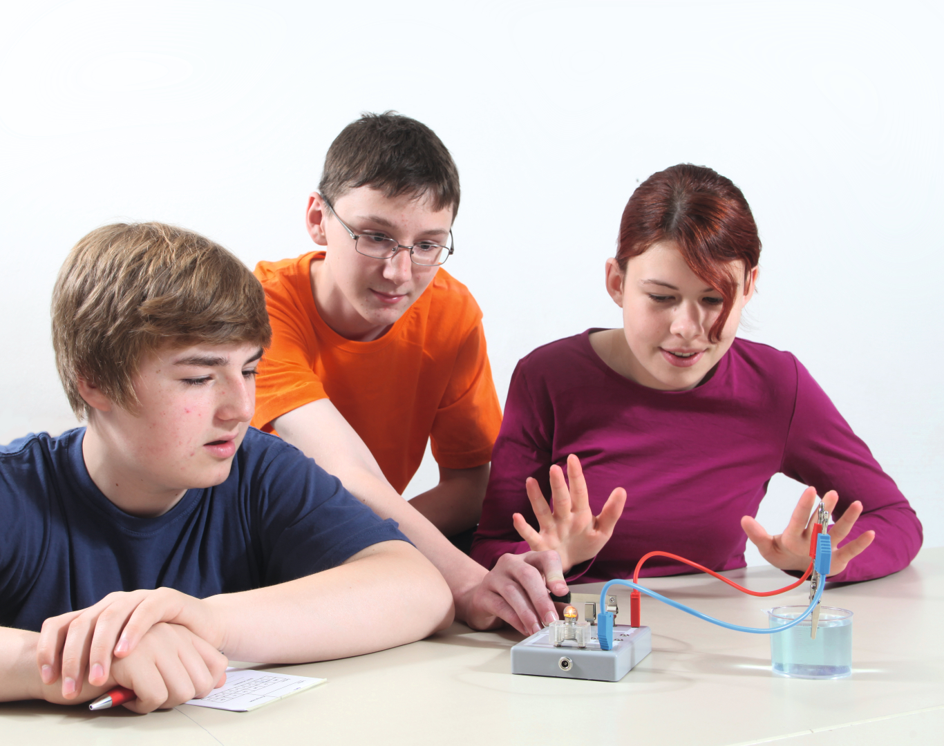 The picture shows three young people working on an experimental device. One of the students is connecting electrical wires while the others watch attentively and take notes to follow the results of the experiment.