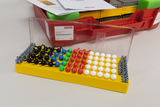 The picture shows a molecular construction kit system with colored balls and rods on a yellow base plate, which is used to illustrate chemical structures. In the background you can see a red storage unit with additional materials and instructions.