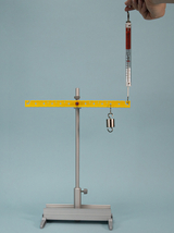 The picture shows an experimental device for carrying out physical measurements. A weight hangs from a hanging scale that is mounted on a yellow horizontal bar, while a handle holds the scale.