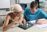The picture shows two students working together on an experiment board. They are experimenting with electrical circuits and have notes and worksheets in front of them.